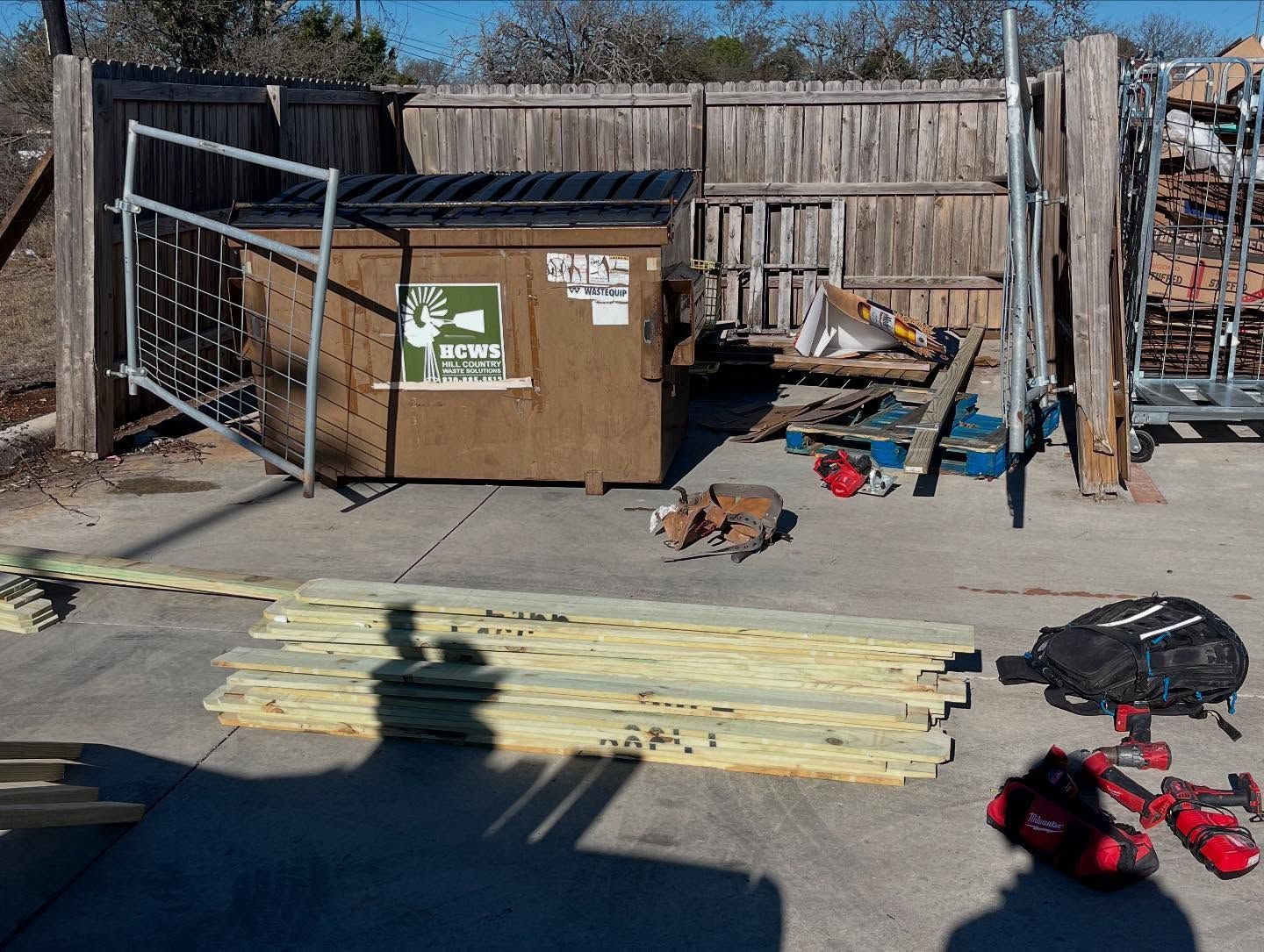 Construction materials and power tools lay on a concrete area near a dumpster and wooden fence.