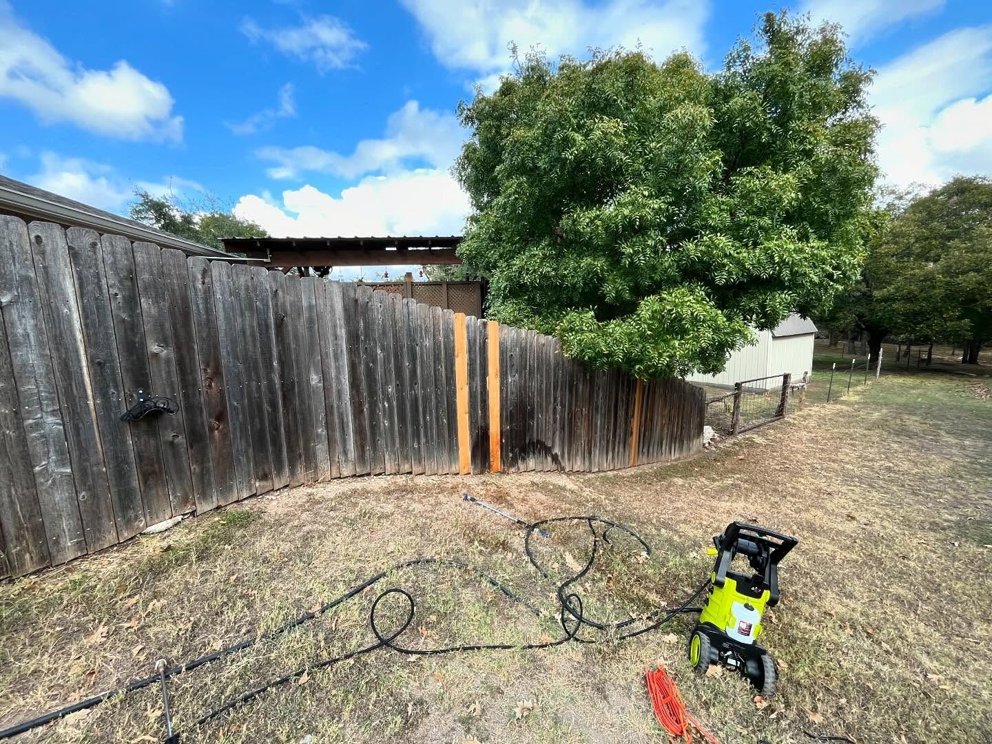 A bright green pressure washer sits on dry grass in front of a long, weathered wooden fence beneath a blue, cloudy sky.