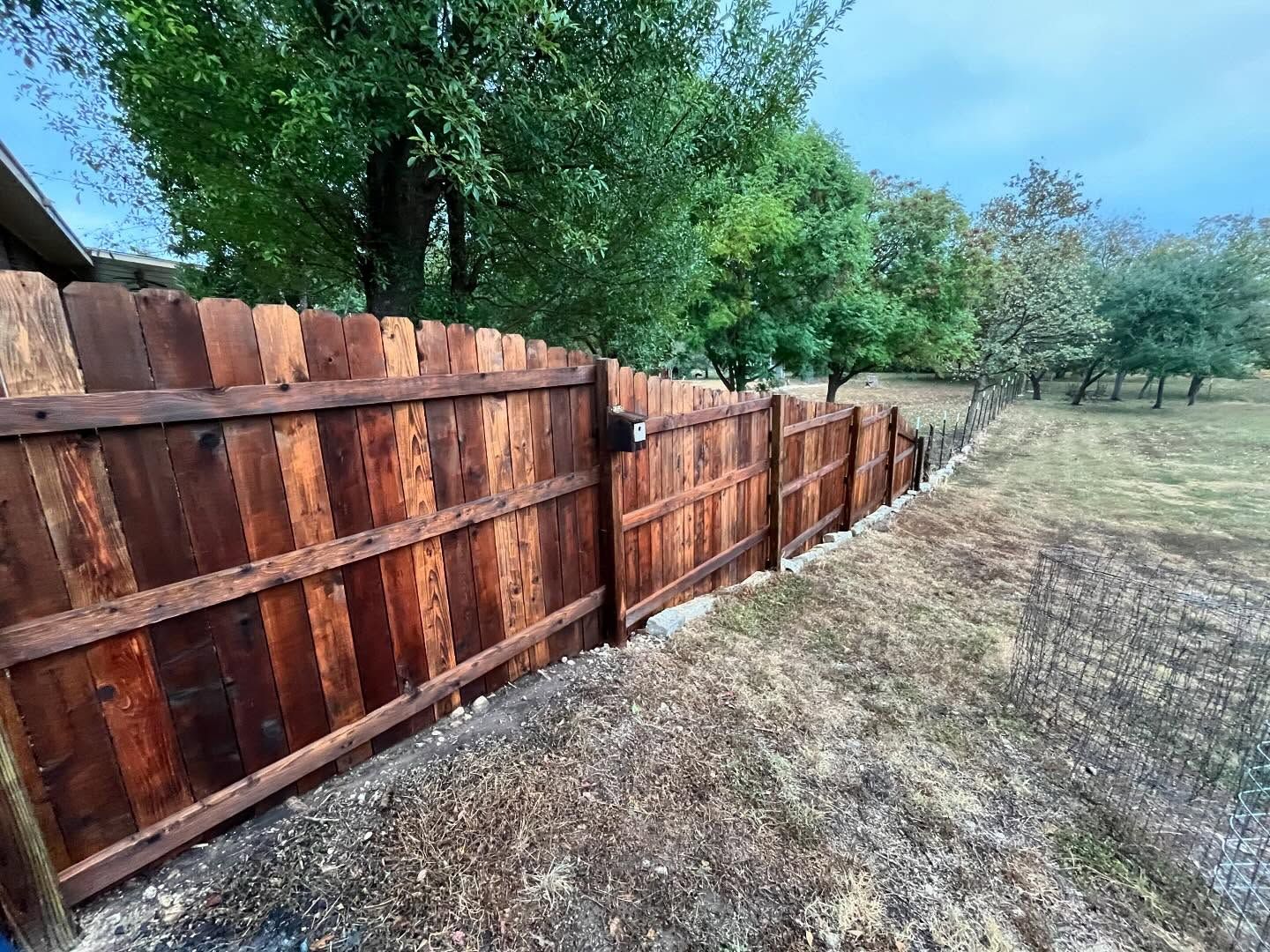 A stained wooden fence stands along a grassy, tree-lined property line under a cloudy sky.