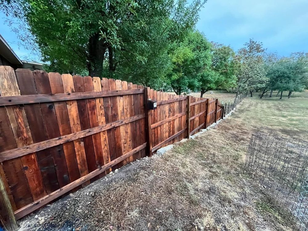 Weathered wooden fence beside a dry grassy yard and green trees under a clear blue sky