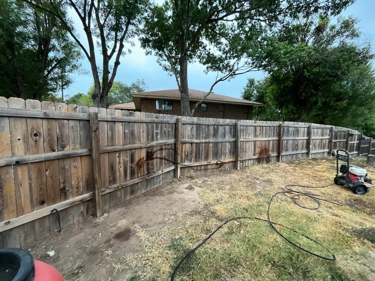 A weathered wooden fence lines a backyard, with a small lawnmower parked on the dry grass in the distance.