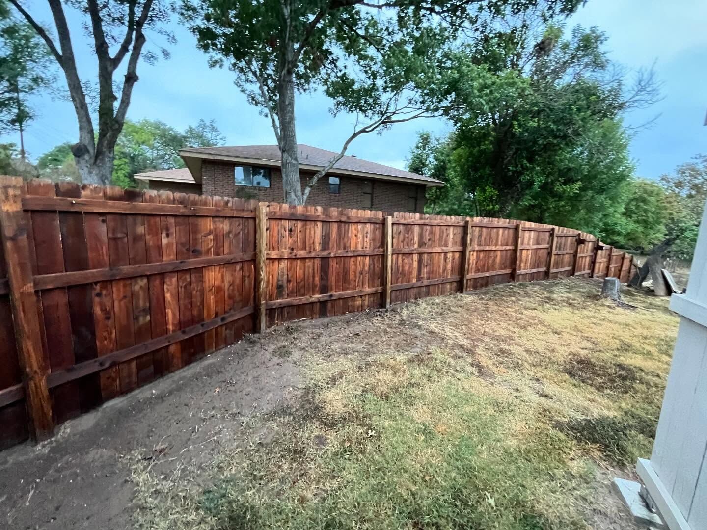 A newly stained, reddish-brown wooden privacy fence runs along a sloped yard in front of a house and trees.