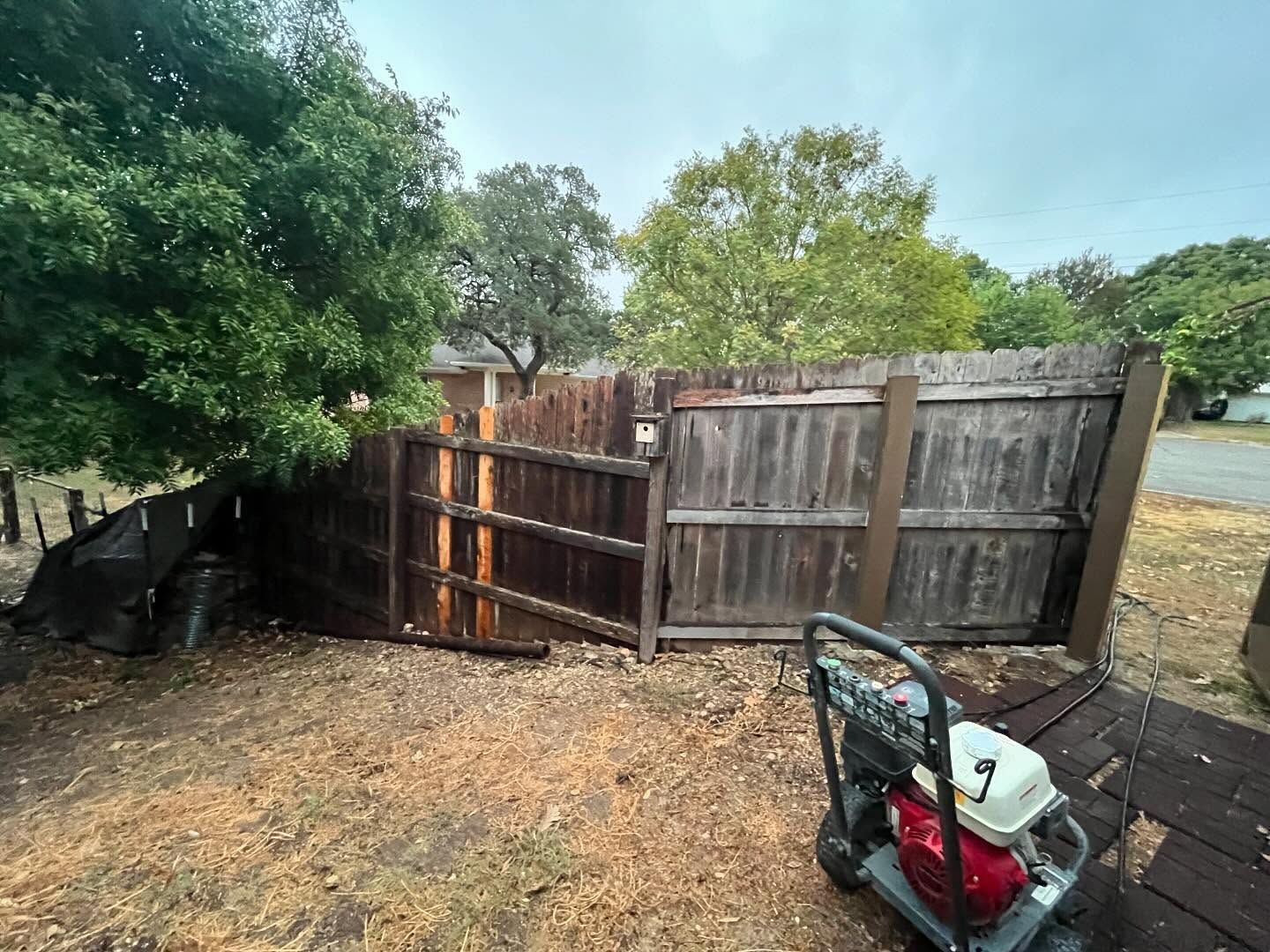 A partially collapsed, weathered wooden fence in a yard with a portable generator in the foreground.