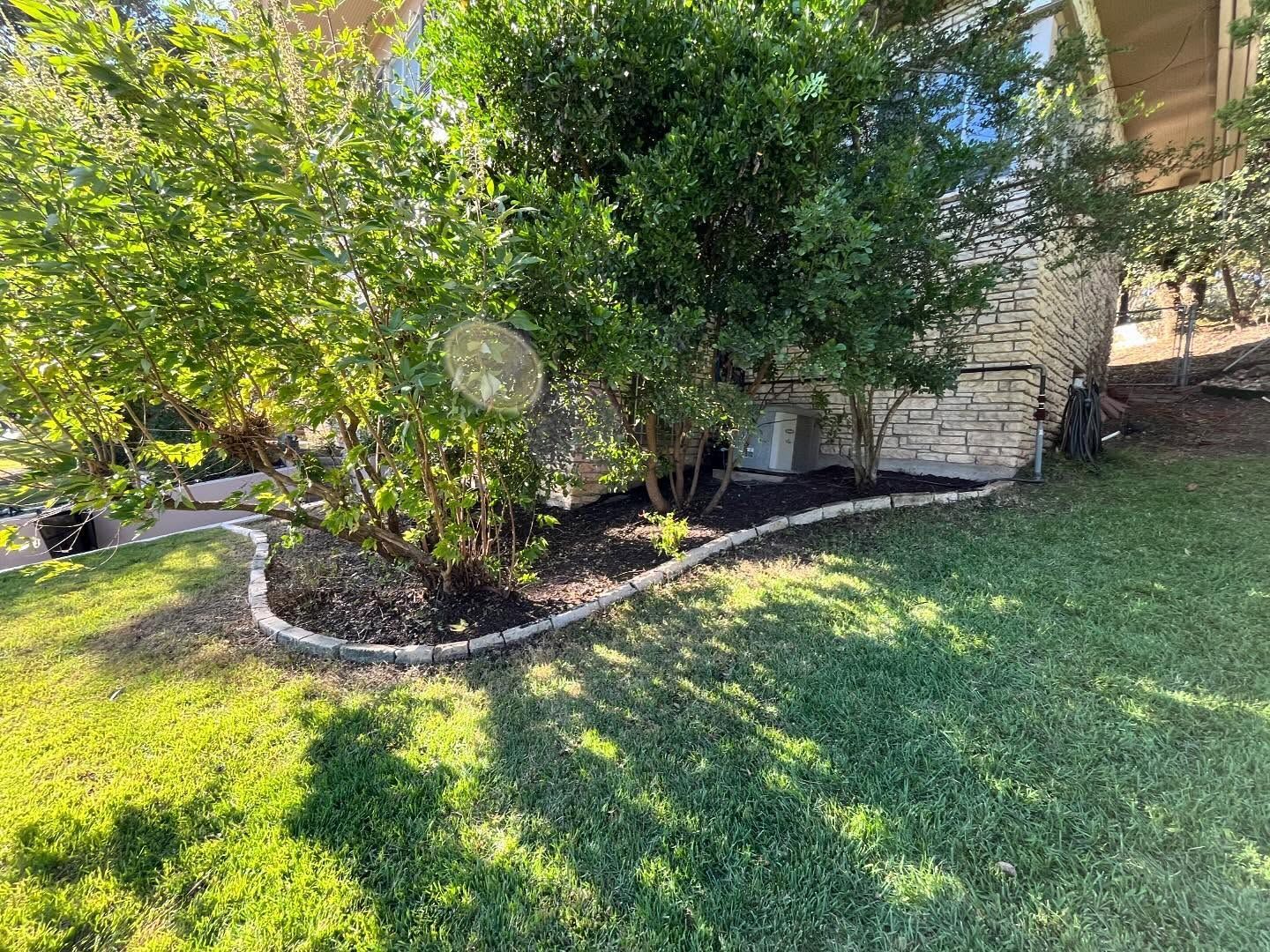 A landscaped garden bed with mulch and stone edging sits in front of a stone house surrounded by green trees and grass.