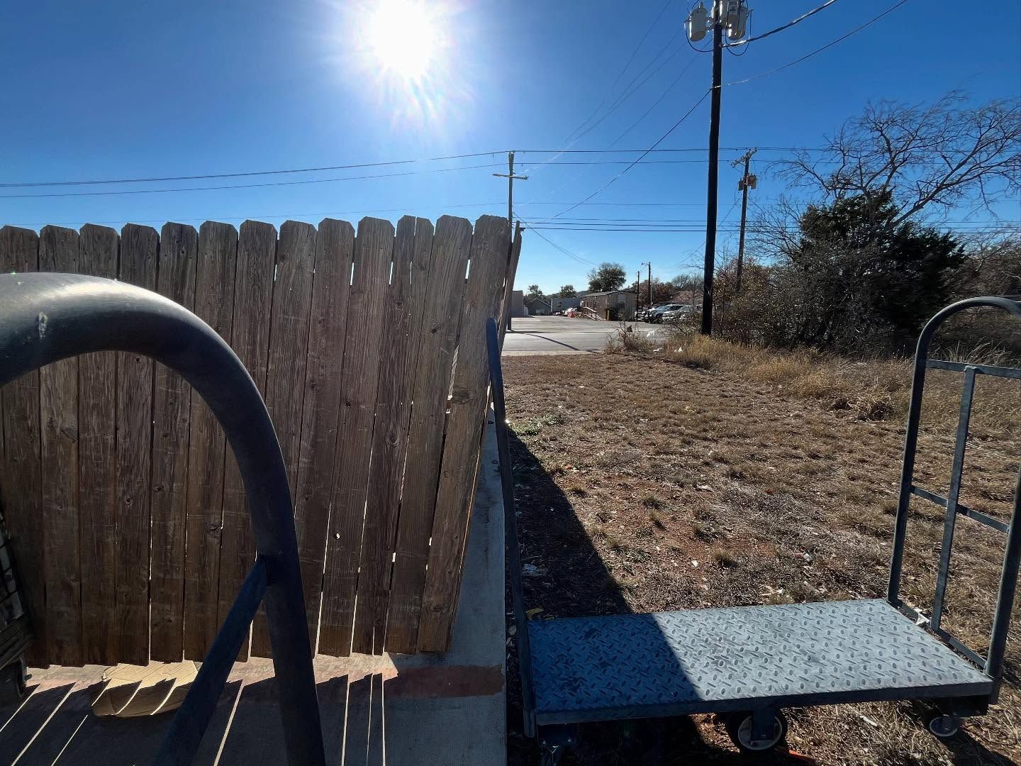 A metal hand truck sits on a dry, grassy patch next to a wooden fence on a sunny day with a clear blue sky.