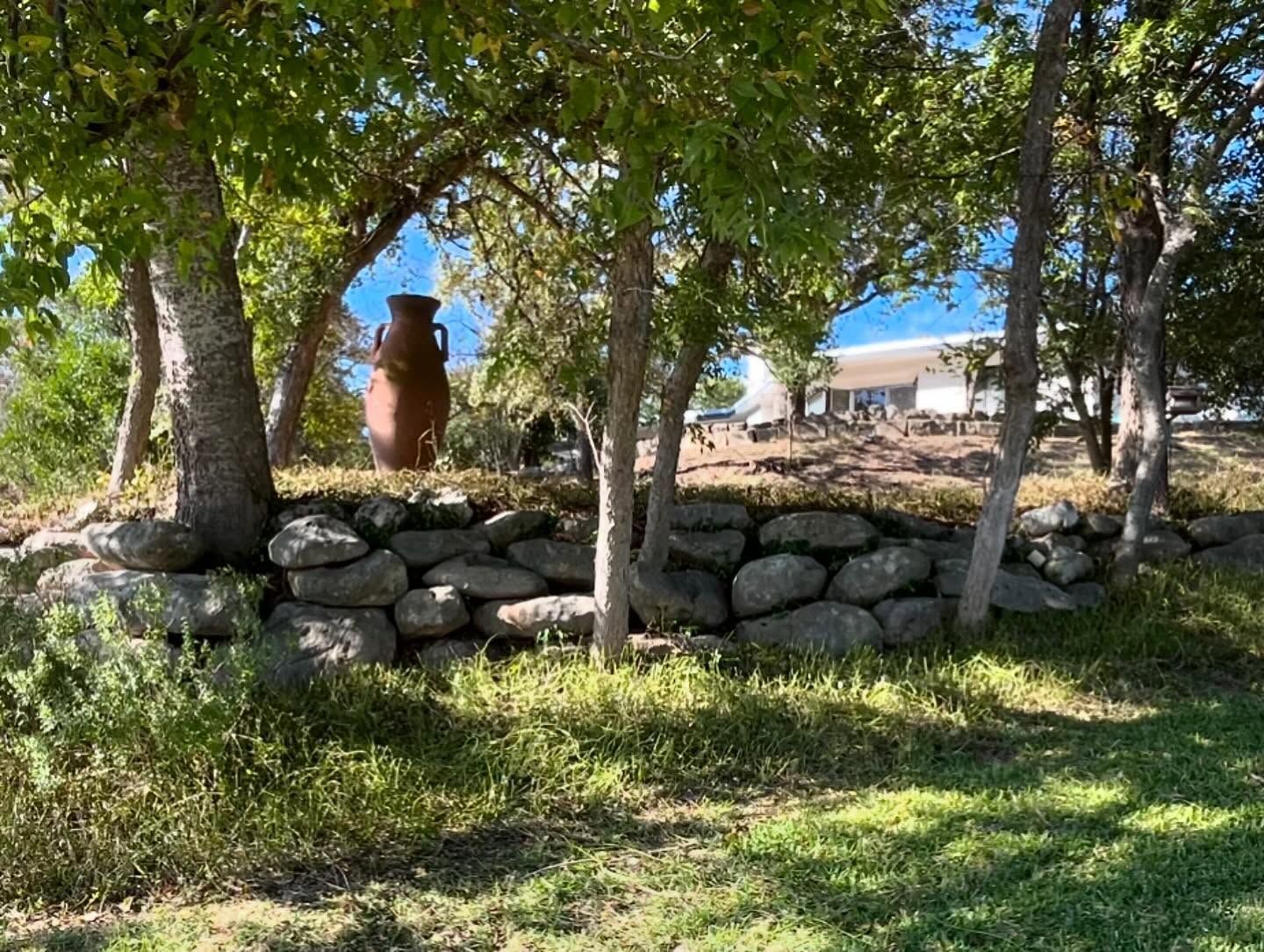 A stone retaining wall built into a grassy hill, shaded by trees and featuring a large decorative brown pottery urn.