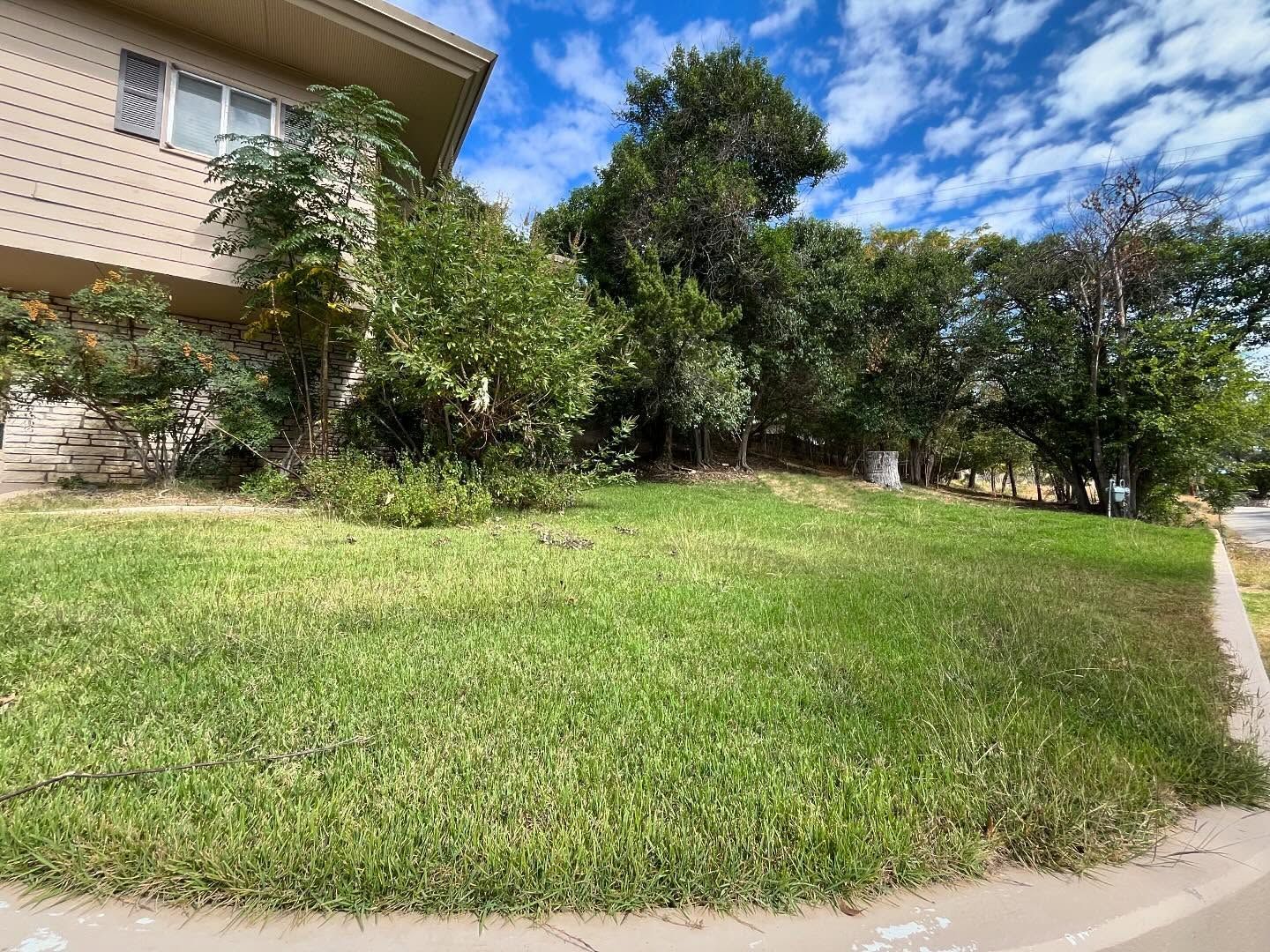 A grassy corner lot slopes upward toward trees next to a beige house under a blue sky with white clouds.
