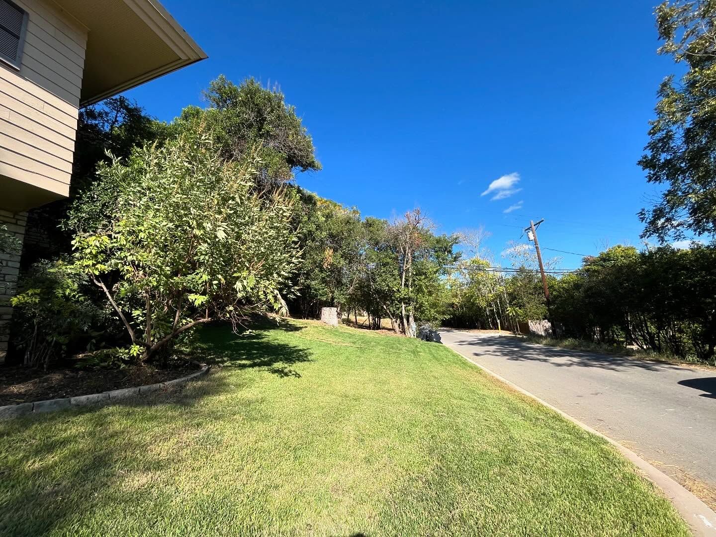 A grassy front lawn slopes up toward a residential building alongside an asphalt road under a clear blue sky.
