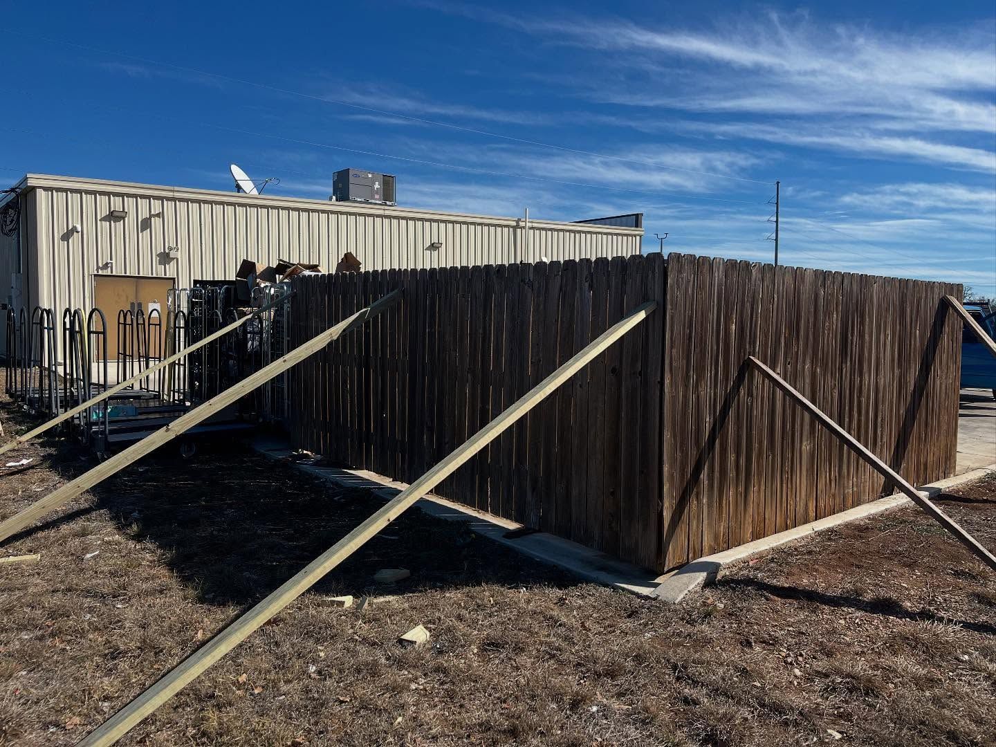 A wooden privacy fence stands in a dirt lot next to a beige building, supported by angled wooden support beams.