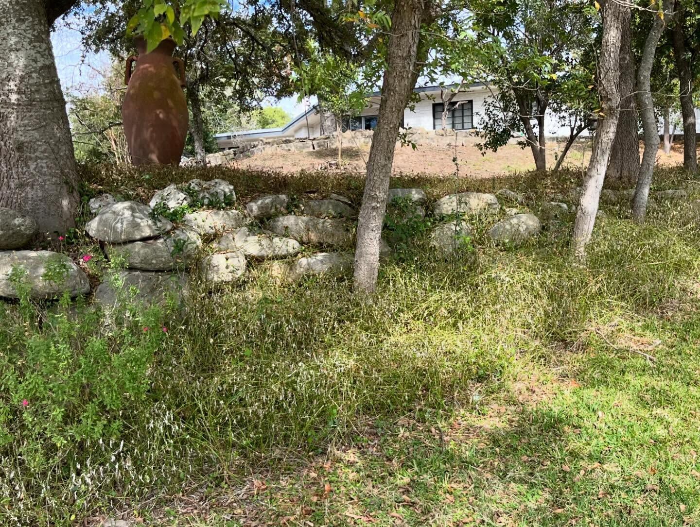 A large stone retaining wall sits under trees, with a house visible in the background atop a grassy, sunlit hill.