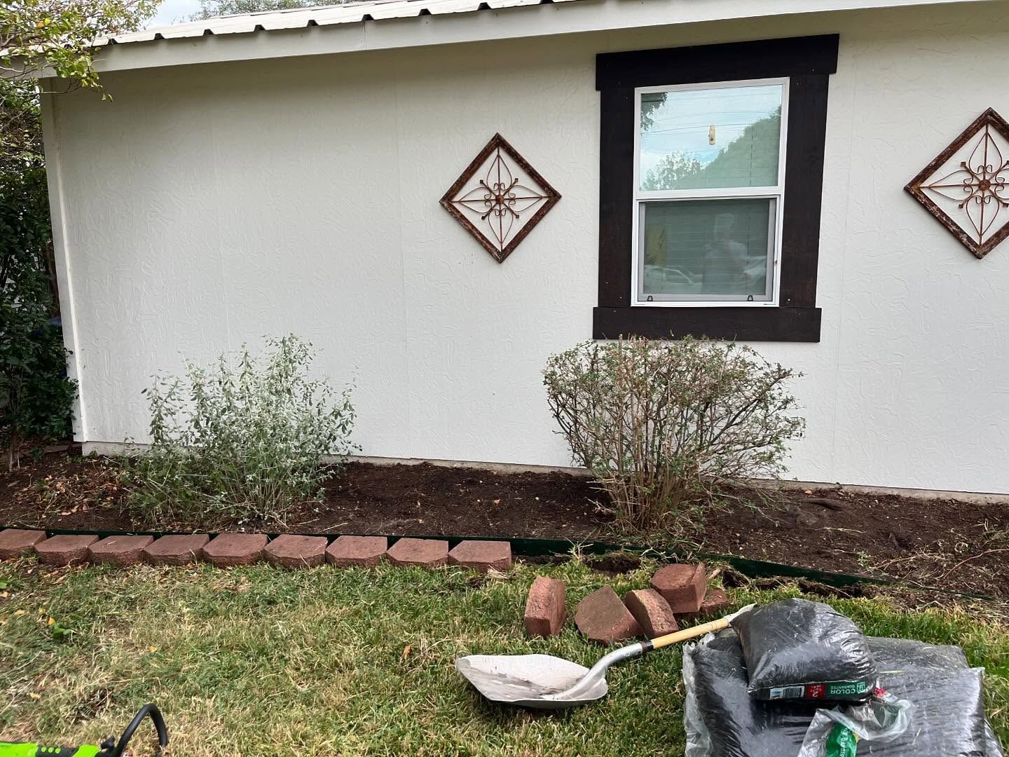 A white wall with a window, two wall decorations, shrubs in a garden bed, and a shovel with bags of mulch on the lawn.