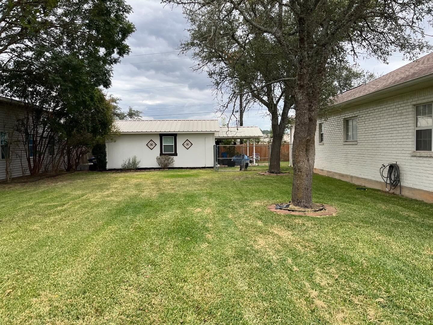 A grass backyard sits between a white detached building and the side of a house, framed by trees under a cloudy sky.