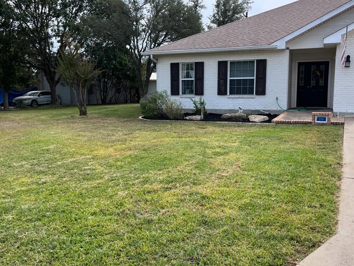 A white brick house with a dark front door and shutters, featuring a large, grassy front yard under a cloudy sky.