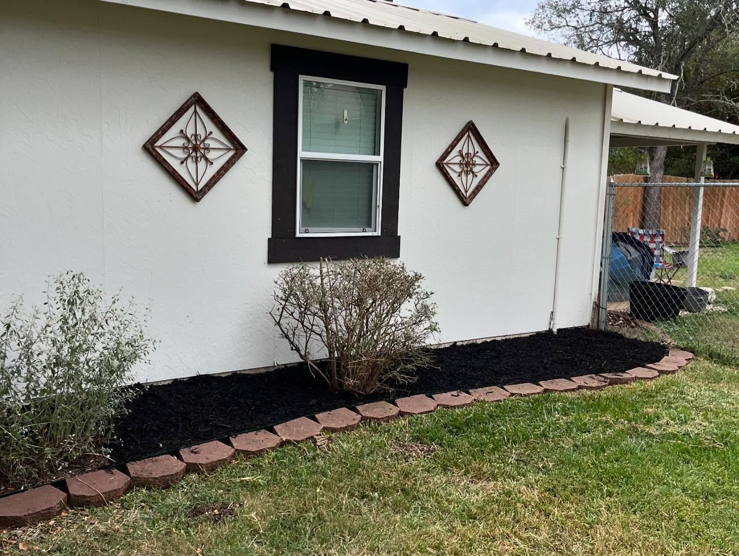 A white exterior wall featuring a window with dark trim, two diamond-shaped wall decorations, and a mulched garden bed.