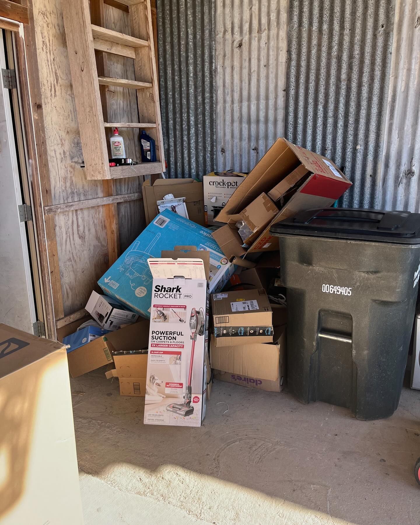 A cluttered storage area featuring cardboard boxes, a vacuum cleaner box, and a dark plastic trash bin against metal walls.