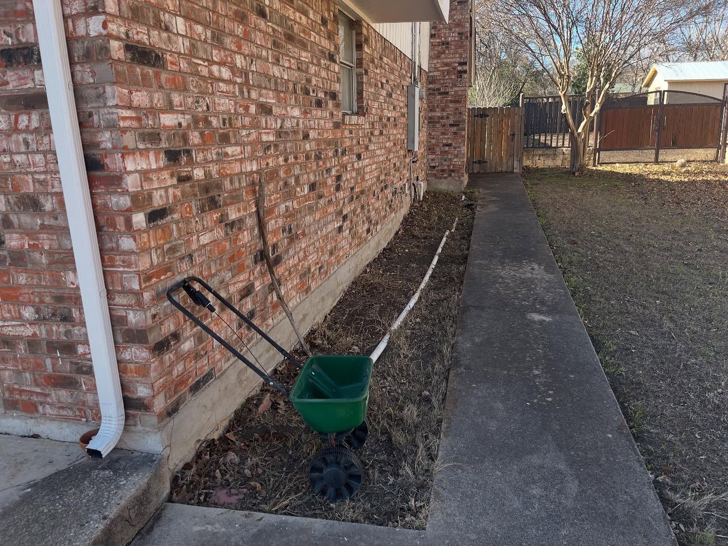A green broadcast spreader sits on a dirt garden bed alongside a brick house and a concrete walkway.