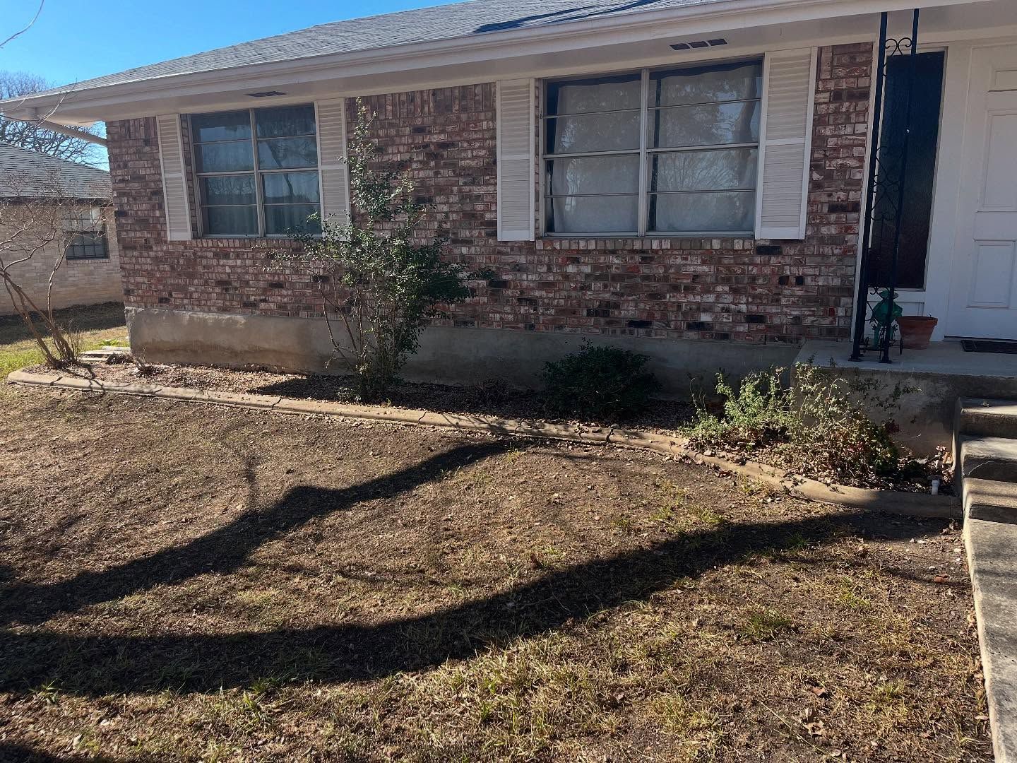 A single-story brick house with white window shutters and a front yard with dry grass and tree shadows on a sunny day.