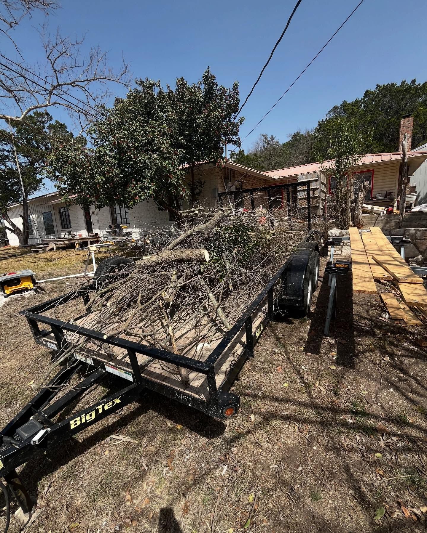 A black utility trailer loaded with tree branches and brush sits on a dirt lot in front of a residential home.