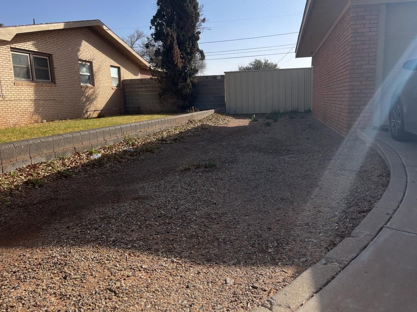 A residential side yard with a gravel ground cover, brick house walls, a concrete driveway, and a tan privacy fence.