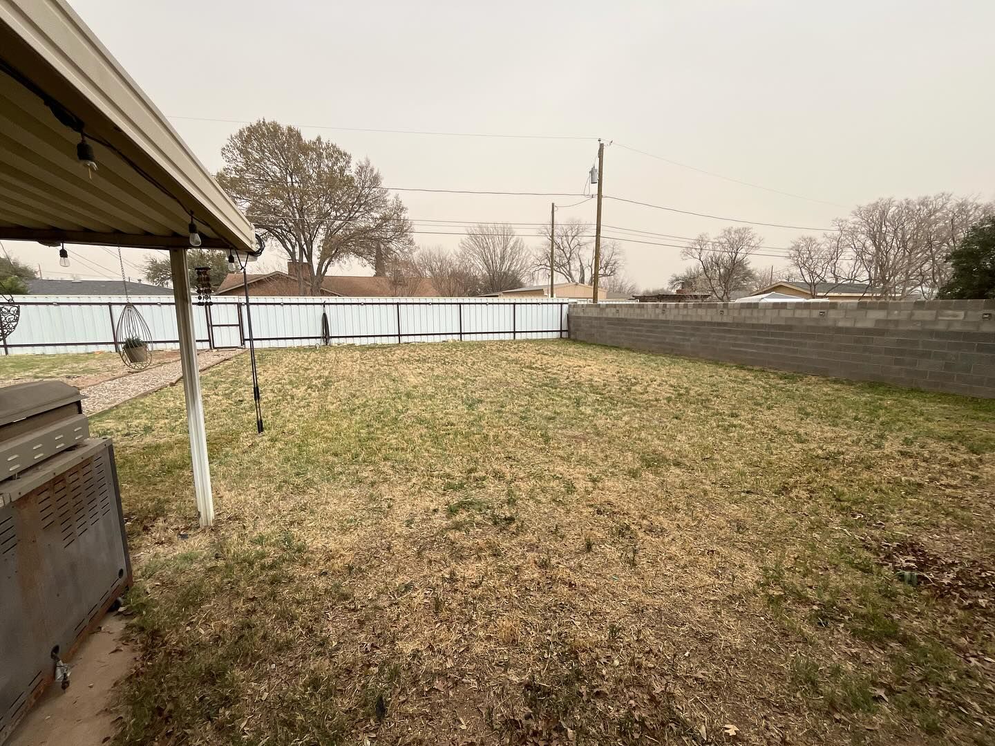 A backyard view with a covered patio, a grassy lawn, a gray stone fence, and trees under an overcast sky.
