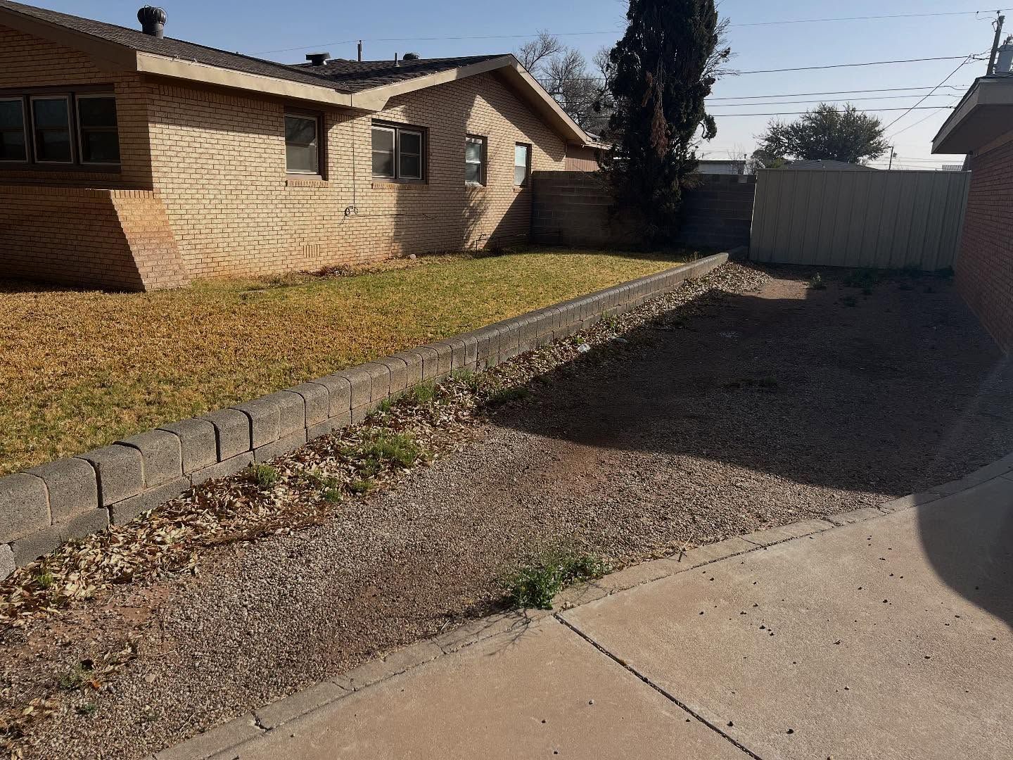 A view of a tan brick house with a low retaining wall separating a grassy yard from a gravel area and concrete patio.