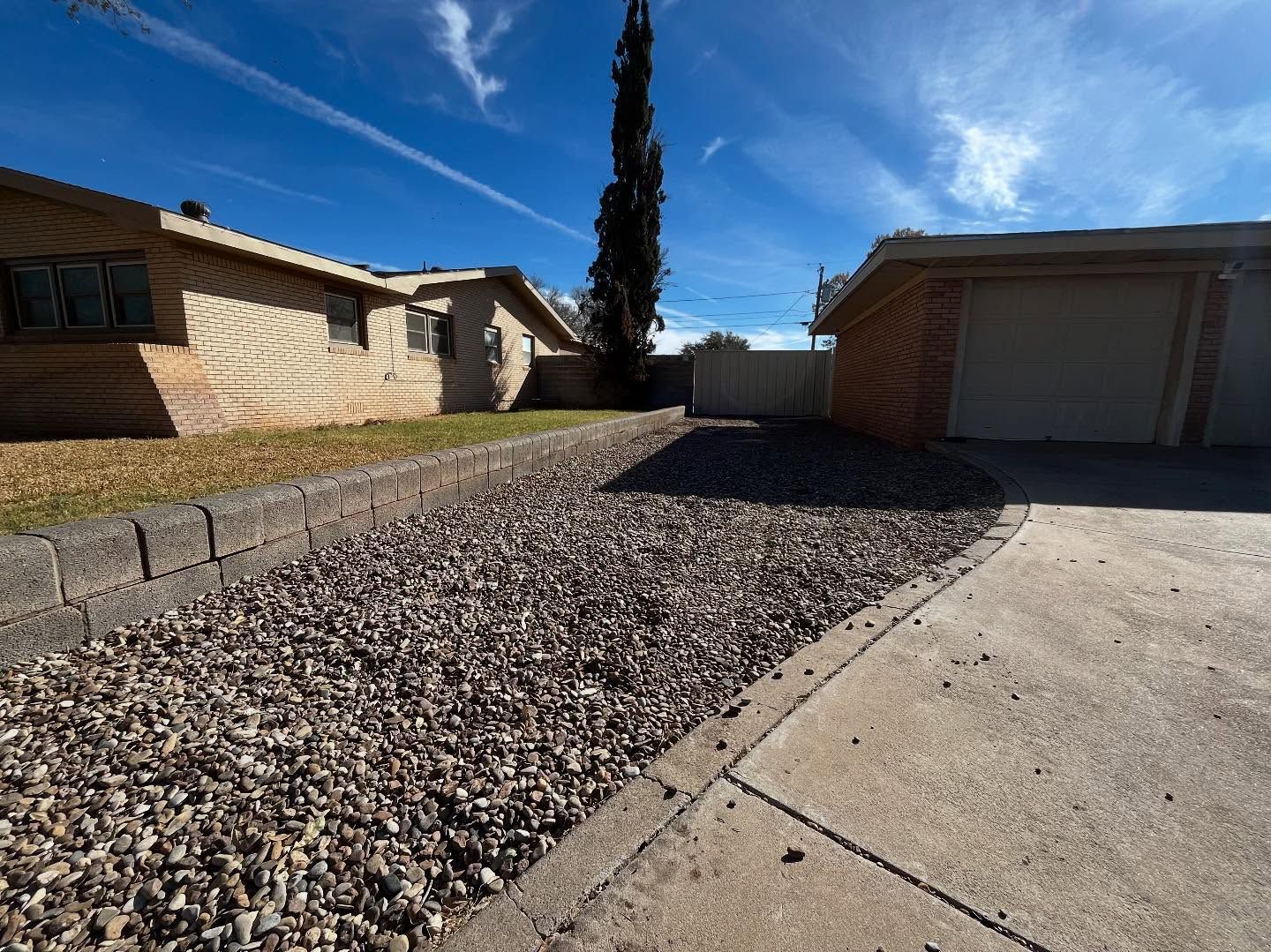 A beige house with a gravel front yard, a concrete driveway, and a tall, thin tree under a bright blue sky.