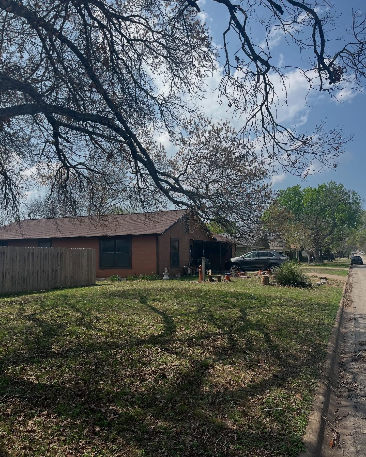 A brick house viewed from a grassy yard, partially shaded by large, bare-branched trees under a bright blue sky.