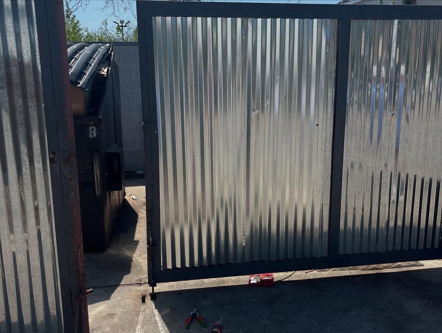A metal gate featuring corrugated panels framed in dark steel, positioned next to a black dumpster on a paved lot.