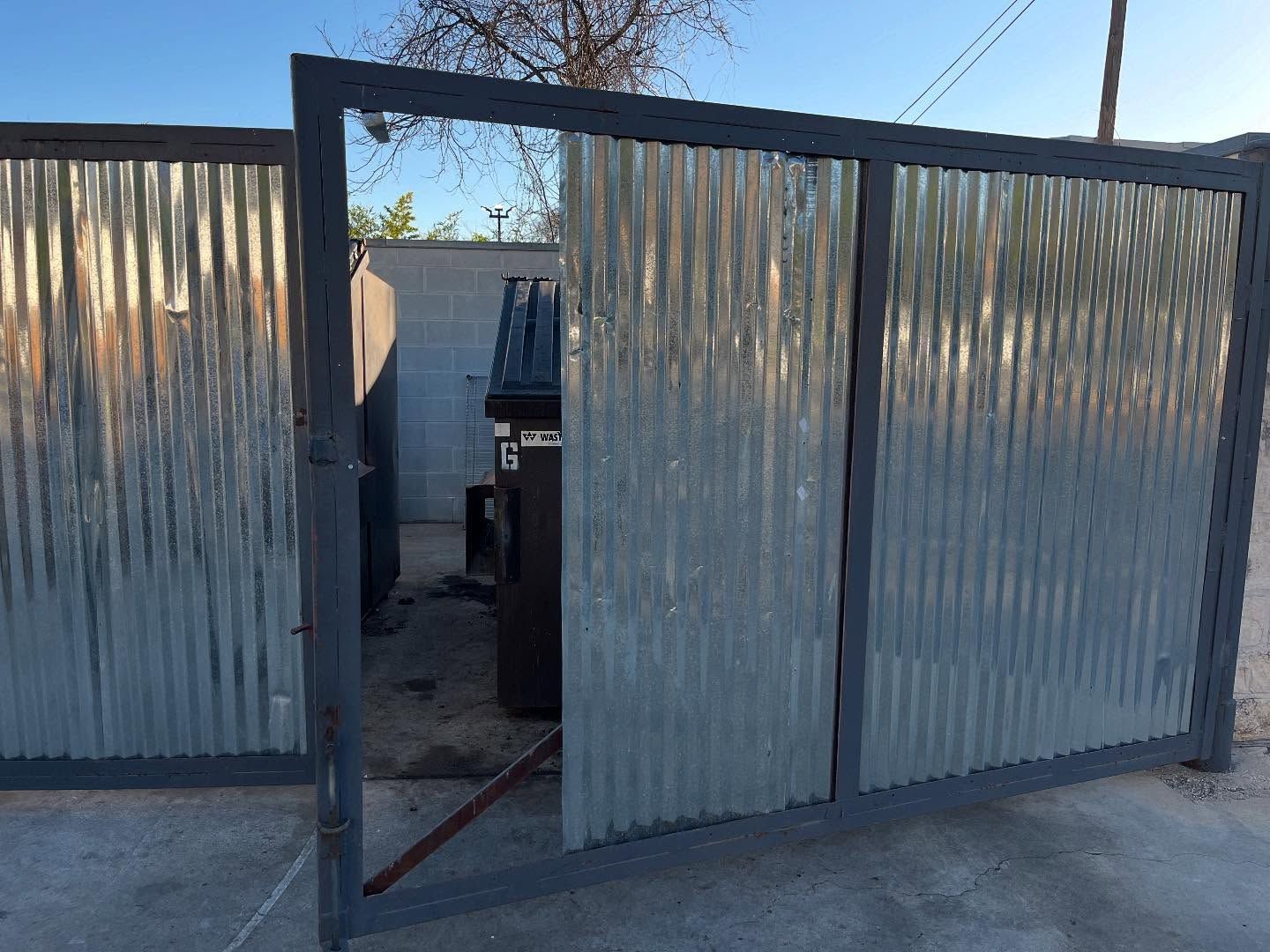 A partially open metal gate made of corrugated steel panels, revealing a dark dumpster inside a fenced enclosure.