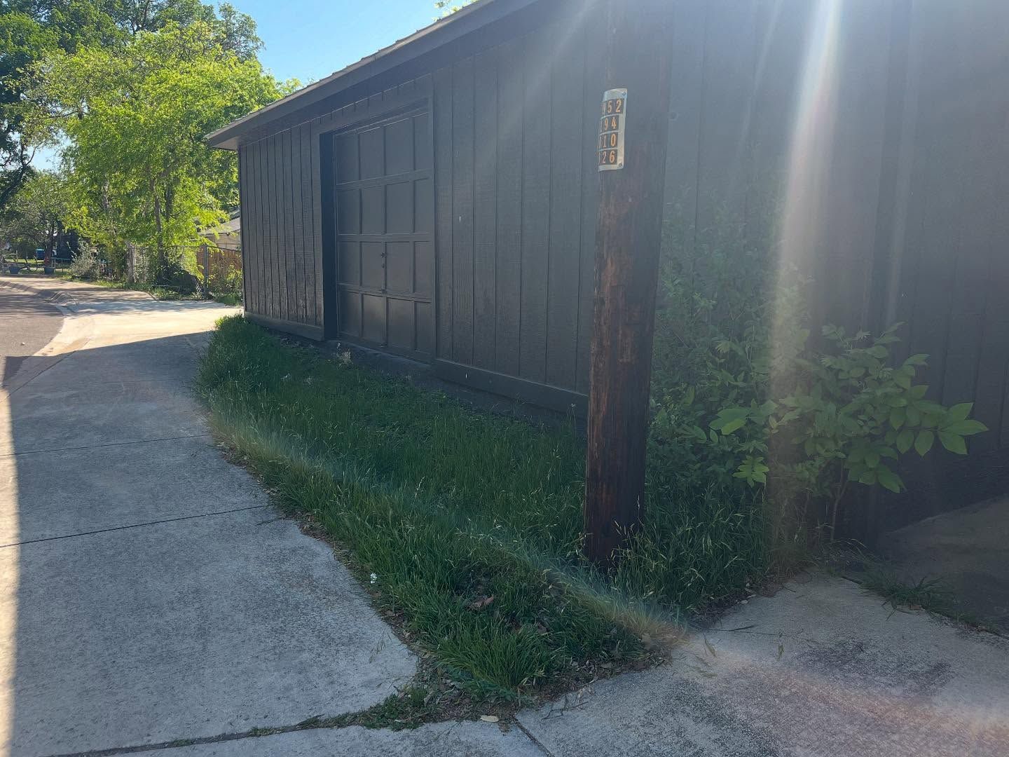A dark brown wooden wall stands beside a concrete walkway with a grassy edge, illuminated by bright sunlight.