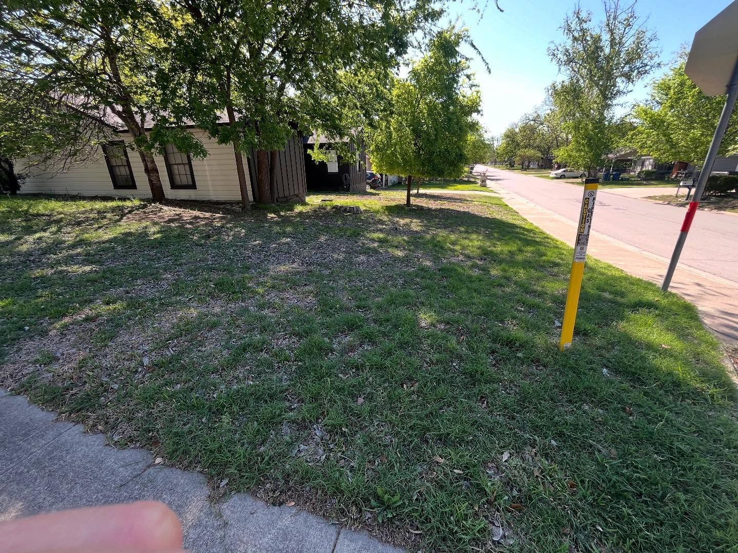 A grassy residential corner lot with a single-story house in the background and a yellow utility post on the sidewalk.