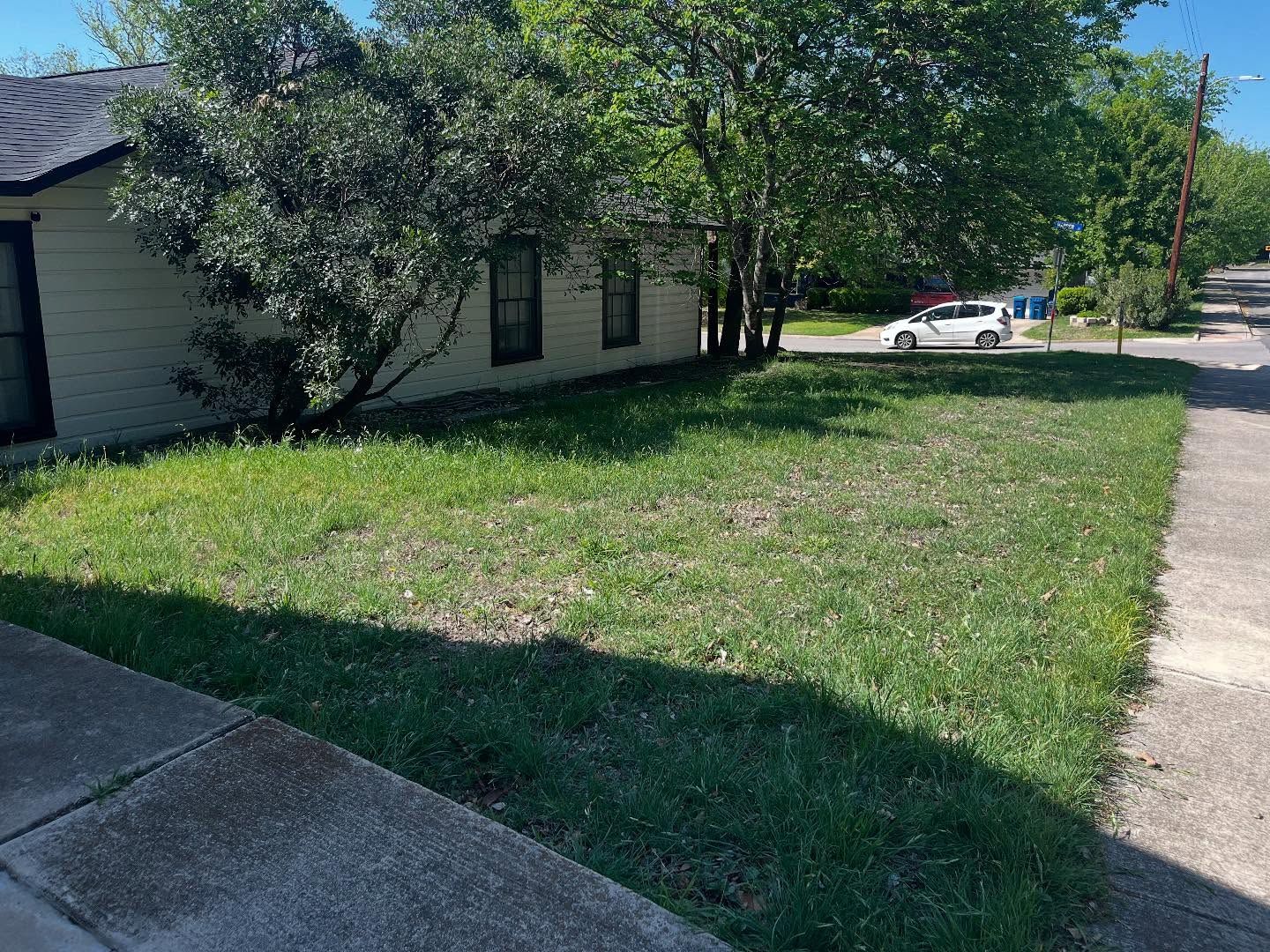 A pale, single-story house sits behind a grassy front lawn next to a sidewalk on a sunny day.
