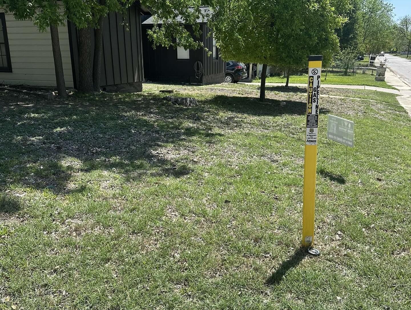 A yellow utility marker post stands in a grassy yard near trees and residential buildings on a sunny day.