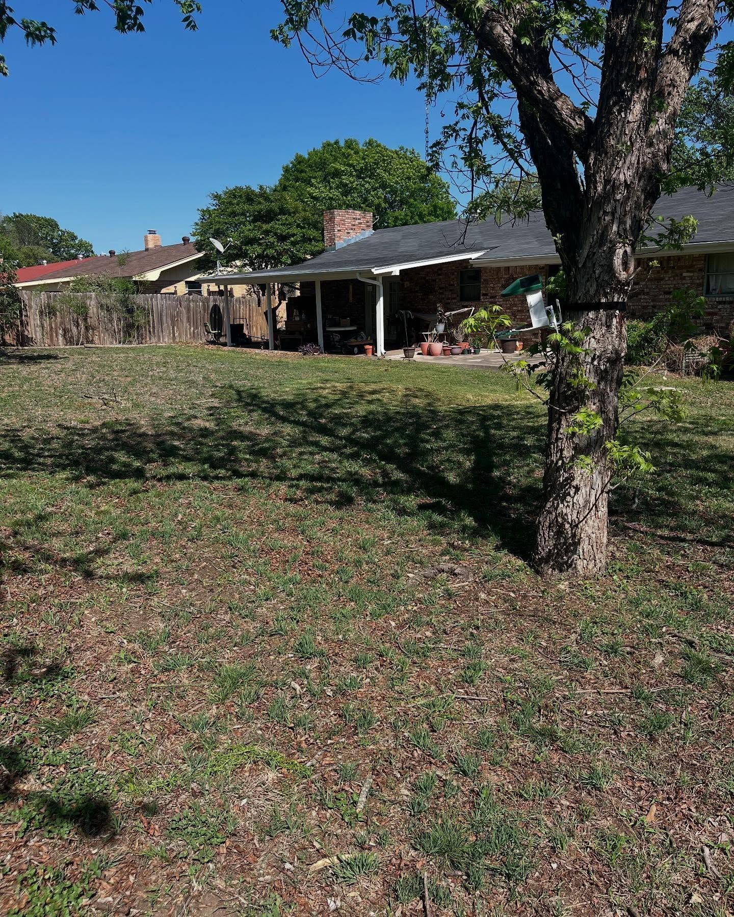 A sunny, grassy backyard leads to a brick house with a covered patio and a brick chimney under a clear blue sky.