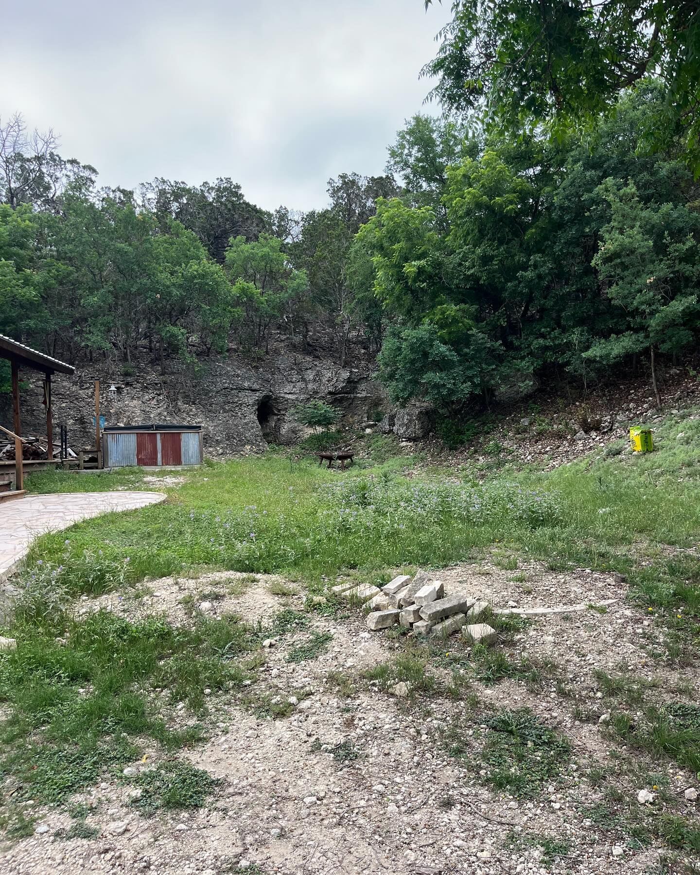 A rural area with a gravel path, a small shed, a rocky stone wall, and dense trees under an overcast sky.