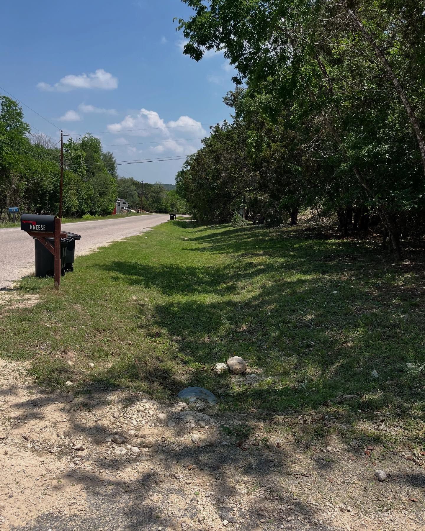 A rural road with a mailbox on the left and a grassy verge bordered by a line of trees on the right under a blue sky.