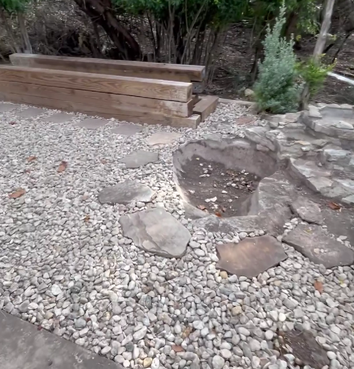 A stone path leads across a gravel yard toward stacked timber garden borders, a small sunken pit, and a young evergreen.