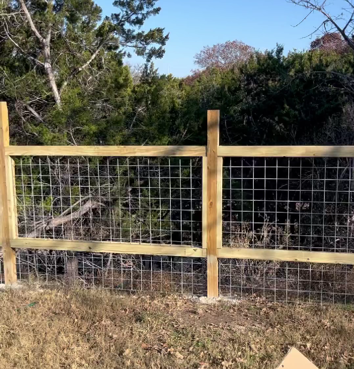 A wooden post-and-rail fence with a wire grid infill installed outdoors in a rural, wooded setting.