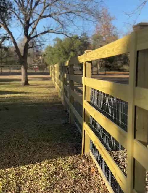 A new wooden three-rail fence with attached wire mesh runs across a grassy yard toward a tree under a clear blue sky.