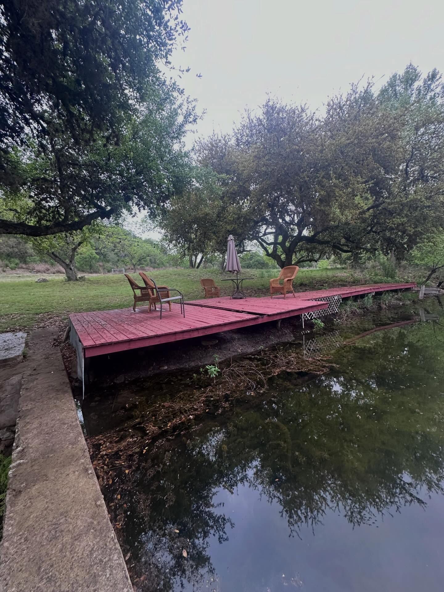 Red wooden dock over calm pond in a green park, with trees and a chair nearby.