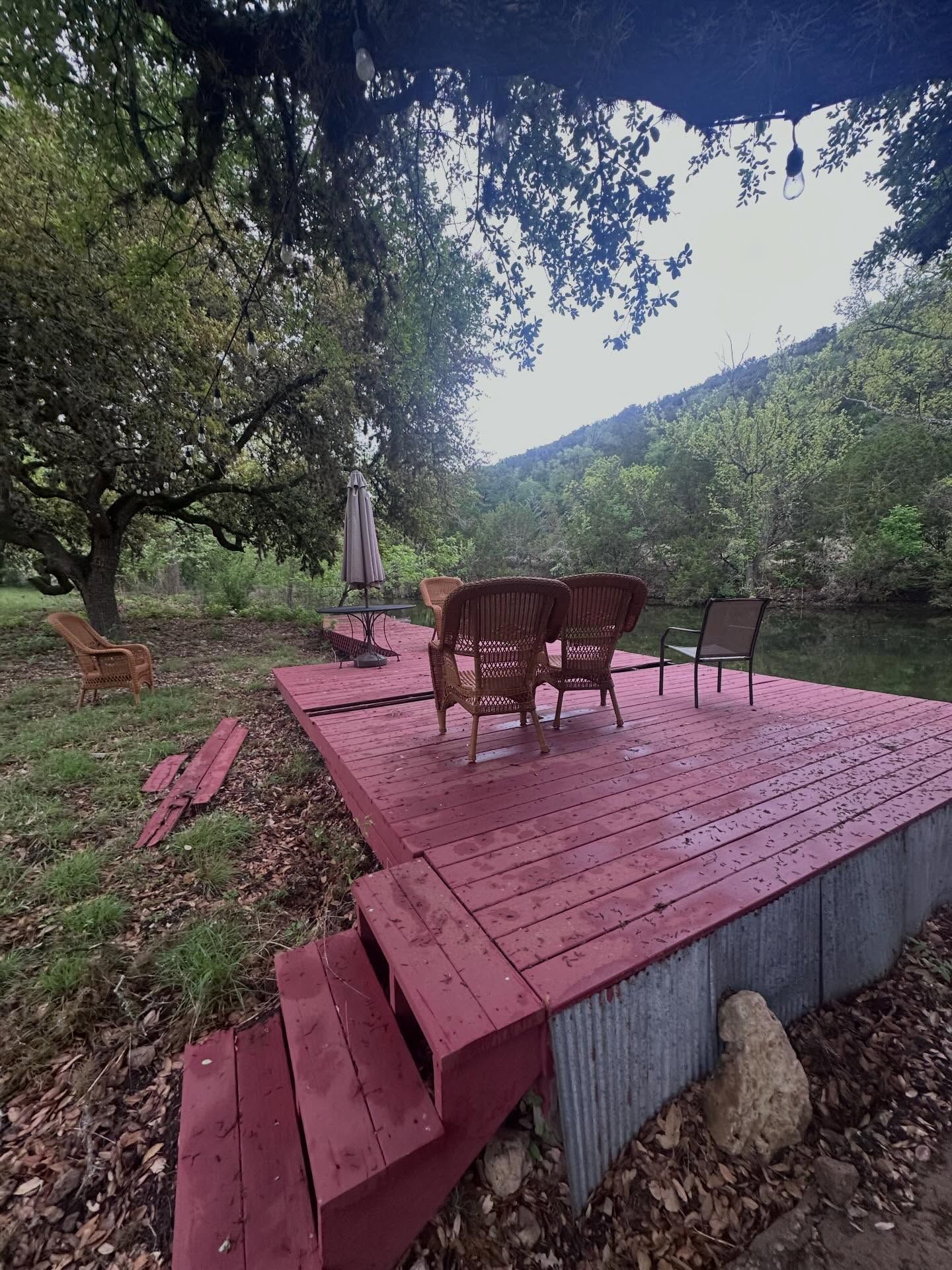 Red wooden deck with chairs and table under trees overlooking a green hillside