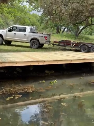 White pickup truck with trailer on a wooden bridge over shallow water in a grassy park.