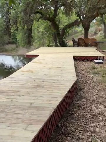 Wooden boardwalk beside a small pond, with trees and outdoor seating in a wooded area.