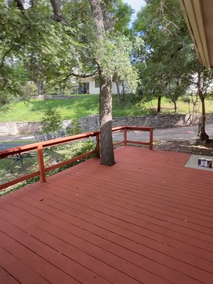 Red wooden deck with railing under trees, overlooking a grassy yard and house.
