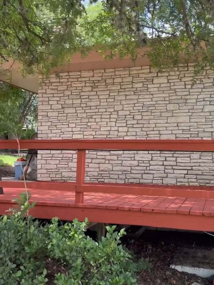Red wooden deck in front of a stone wall, shaded by tree branches and surrounded by greenery