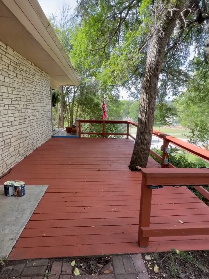 Red wooden deck beside stone house, with trees and railing overlooking a grassy park.