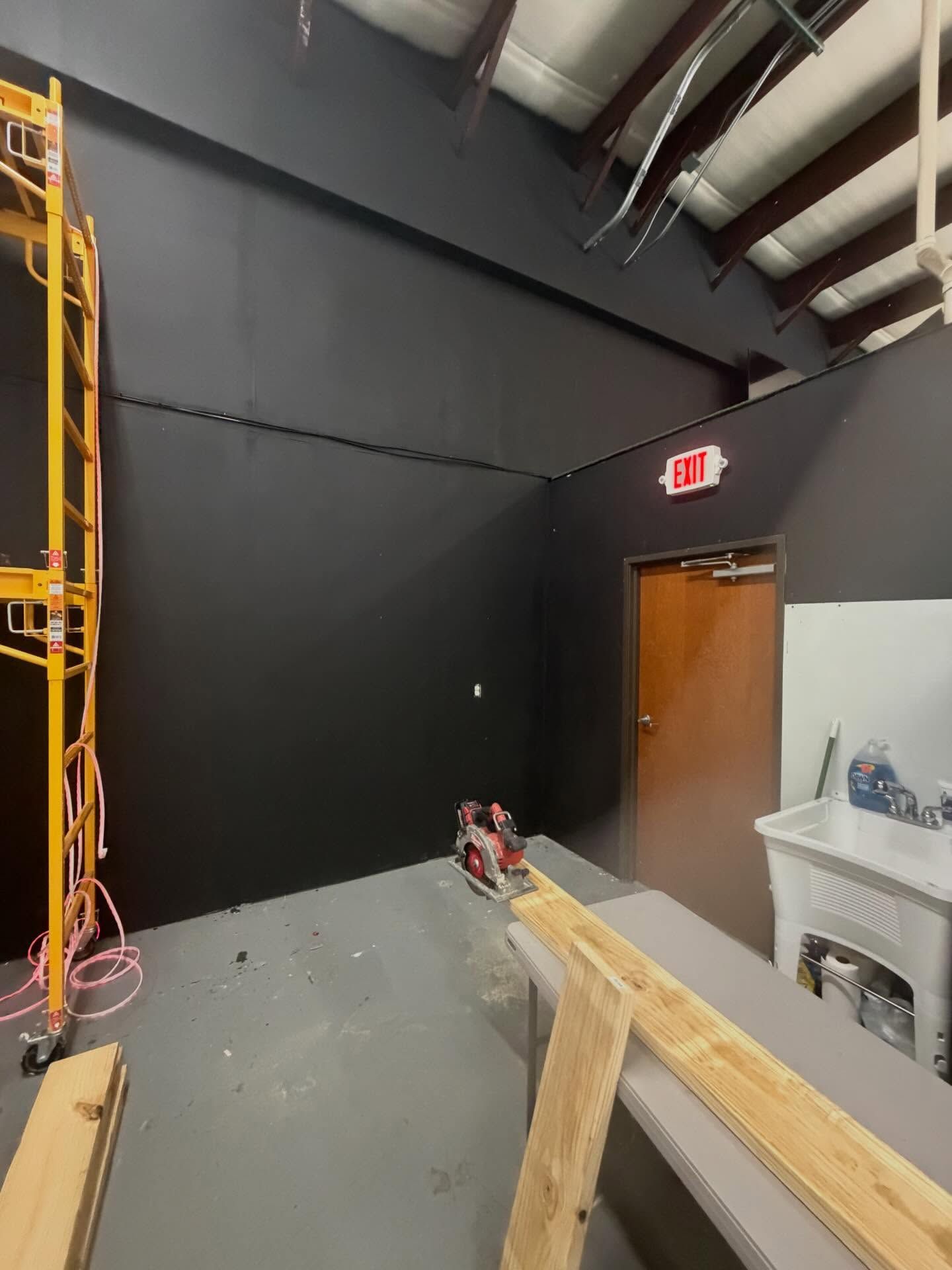 Empty industrial room with black walls, exposed pipes, a door, and a wooden railing in the foreground
