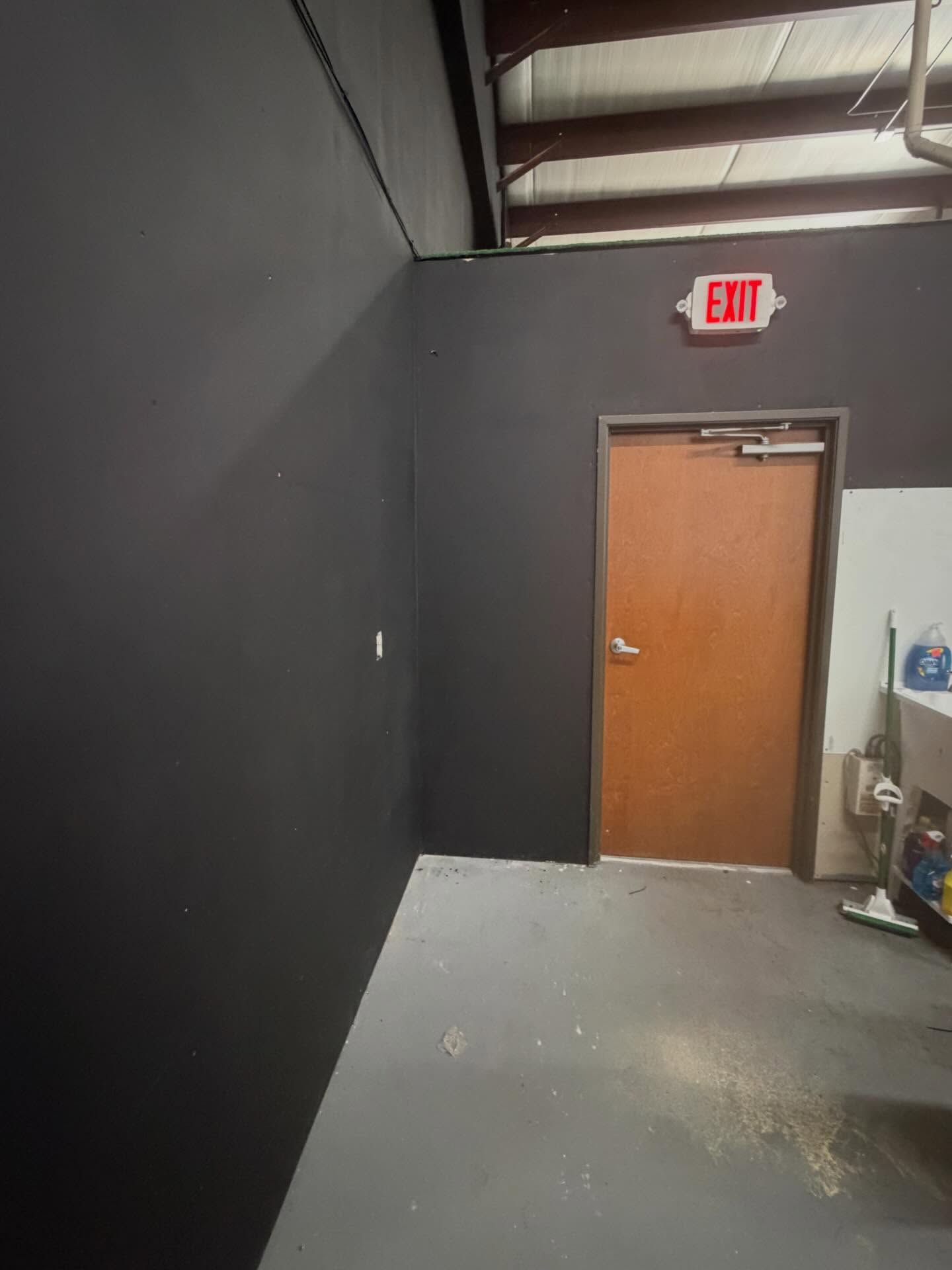 Industrial hallway with black walls, gray floor, and a wooden exit door under a red EXIT sign.