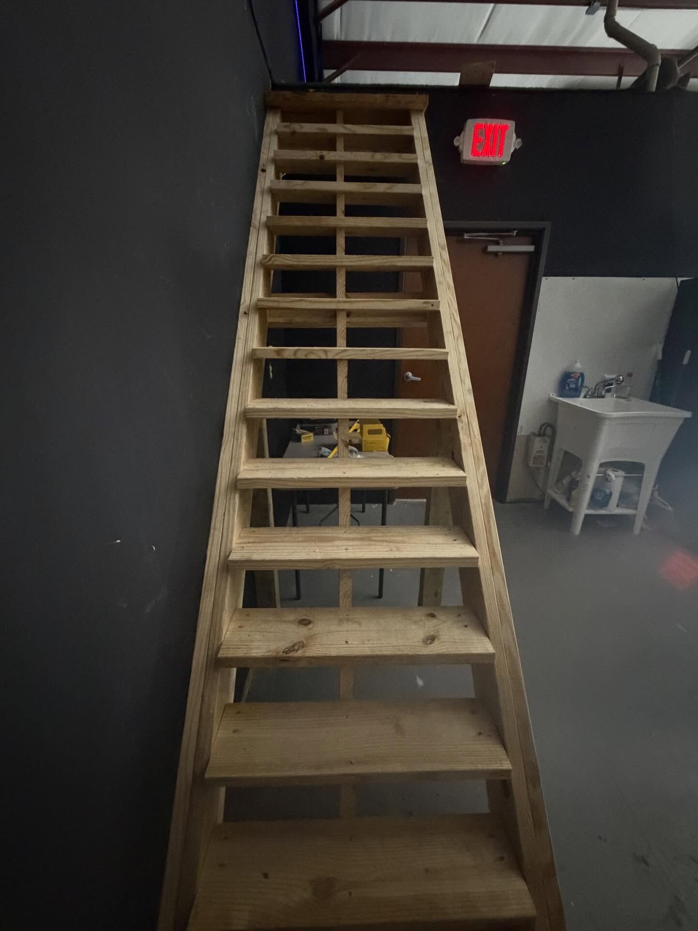 Steep wooden staircase in an industrial room with a red EXIT sign and a small desk in the background
