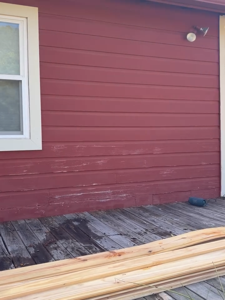 Red house exterior wall with a window, deck boards stacked below, and a small light fixture above.